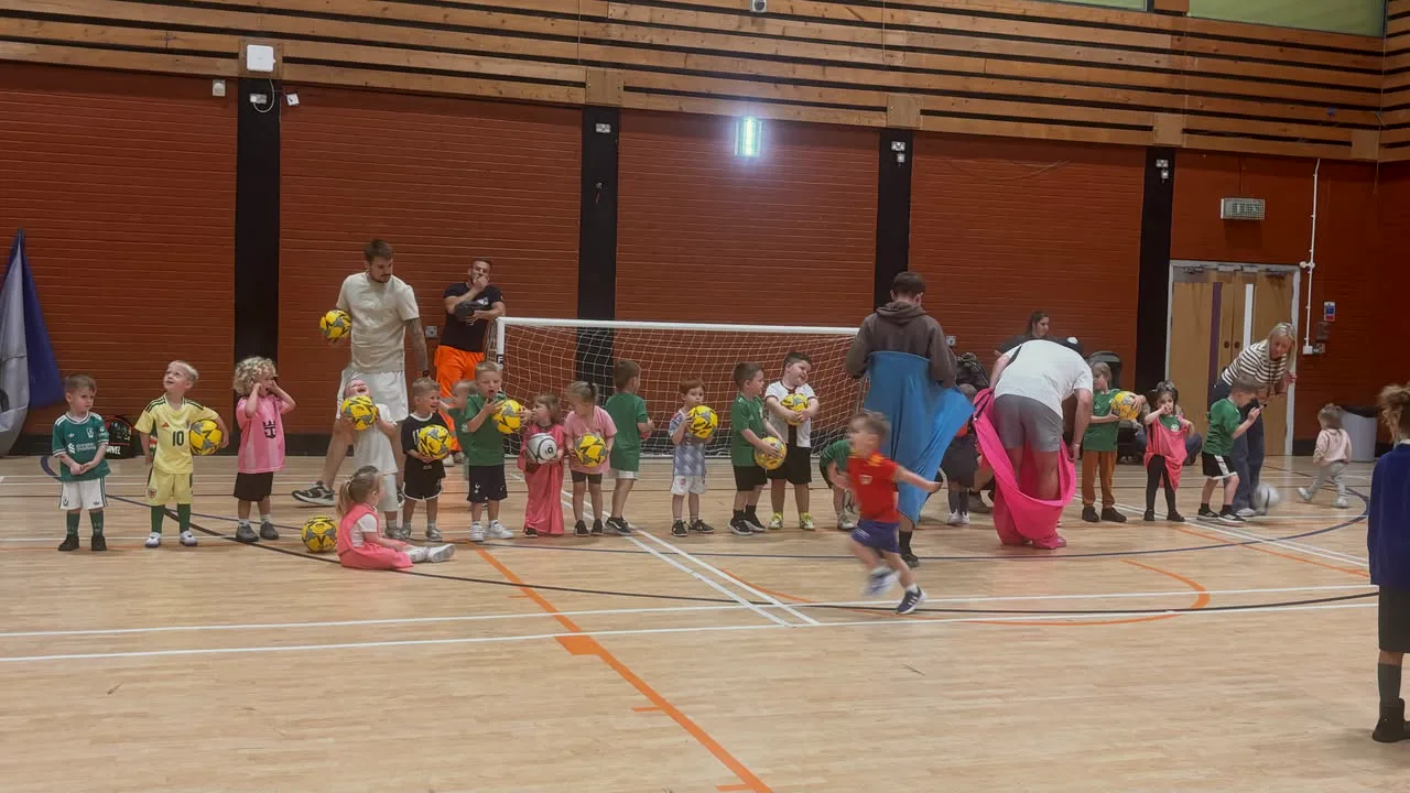A Mini Dragons session in a sports hall: children lined up with footballs while coaches set up the next drill.